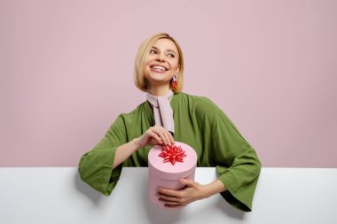 Çekici bir genç kadın elinde bir hediye kutusu ve pembe arka planda gülümsüyor.