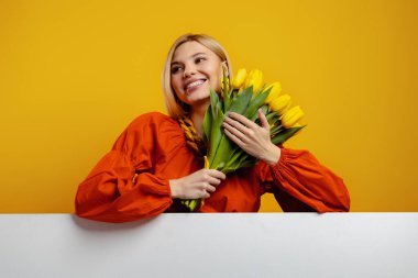 Bir sürü lale tutan ve sarı arka planda gülümseyen çekici genç bir kadın.