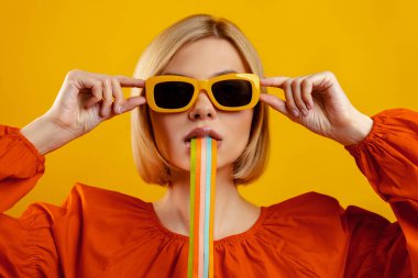 Ağzından sarkan renkli çizgileri olan şık bir genç kadın sarı arka planda duruyor.