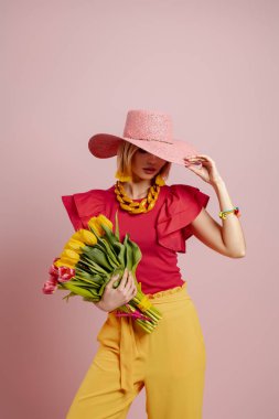 Zarif şapkalı güzel bir kadın pembe arka planda bir demet lale tutuyor.