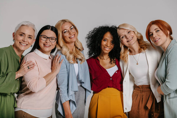 Multi-ethnic group of beautiful mature women bonding and smiling against grey background