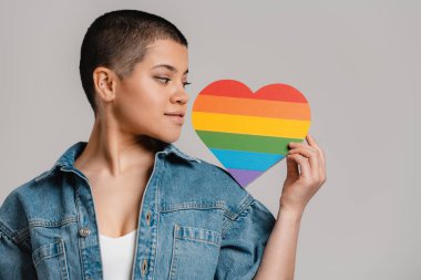 Confident young short hair woman holding heart shaped rainbow flag against grey background clipart