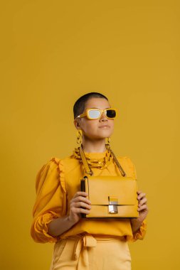 Şık gözlüklü, kısa saçlı, şık bir kadın sarı arka planda moda bir çanta taşıyor.