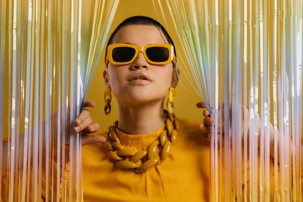 时髦的短发女人 戴着时髦的眼镜 在黄色的背景下 透过彩色的窗帘看 — 图库照片