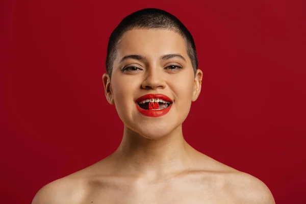 Playful young shaved head woman holding plastic letters in mouth against red background