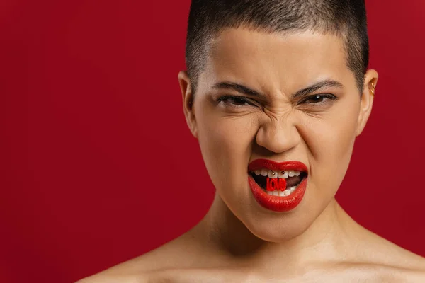 Young shaved head woman holding plastic letters in mouth and grimacing against red background