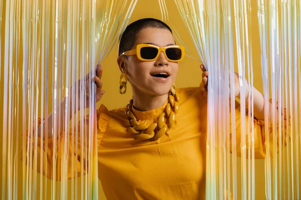 Возбужденная Женщина Модных Очках Смотрит Через Красочный Занавес Желтом Фоне — стоковое фото