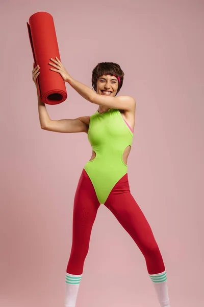 背景色を背景に運動マットを運ぶレトロスタイルのスポーツウェアで幸せなフィット女性 — ストック写真