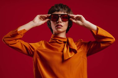 Çekici kısa saçlı kadının portresi gözlüğünü kırmızı arka plana göre ayarlıyor.