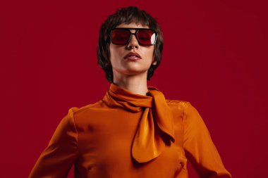 Kırmızı arka planda duran gözlüklü çekici kısa saçlı kadının portresi.