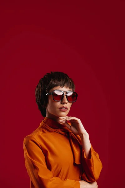 迷人的年轻短发女人 戴着时髦的眼镜 手挽着下巴 背对着红色背景 — 图库照片
