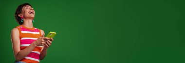 Mutlu genç bir kadın elinde akıllı bir telefonla yeşil arka planda dururken gülümsüyor.