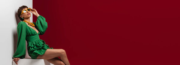 Attractive young short hair woman in elegant dress adjusting her eyeglasses against red background