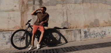 Dışarıda beton bir duvara yaslanmış bisikletine yaslanmış yakışıklı genç bir adam.