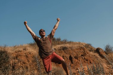 Sırt çantalı mutlu erkek maceracı dağlarda yürürken el kol hareketi yapıyor.