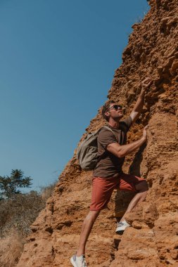 Sırt çantalı kendine güvenen erkek maceracı kayaya tırmanırken tutunuyor.