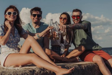 Bir grup mutlu genç arkadaş, deniz kenarında kaygısızca vakit geçirirken birayla kadeh kaldırıyor.