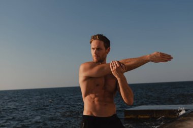 Kendine güvenen erkek sporcu, arka planda deniz varken dışarıda esneme egzersizleri yapıyor.