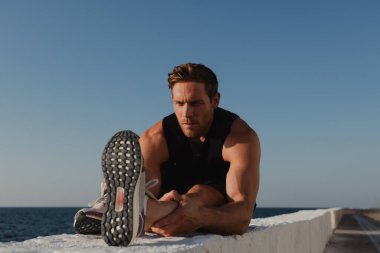 Spor kıyafetli yakışıklı genç erkek sporcu deniz kenarında koşmadan önce esneme egzersizleri yapıyor.