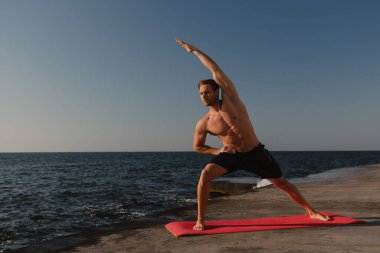 Kendine güveni tam genç bir adam, arka planda deniz varken dışarıda esneme egzersizleri yapıyor.