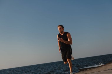 Spor elbiseli kendine güvenen atletik adam arka planda deniz varken açık havada koşuyor.