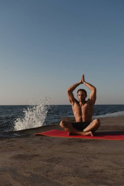 Deniz kenarında meditasyon yaparken egzersiz minderinde oturan yakışıklı genç adam.