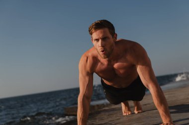 Arka planda deniz varken dışarıda egzersiz yaparken şınav çeken güçlü, genç bir adam.