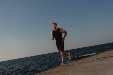 Arka planda deniz varken rıhtımda koşan spor elbiseli kendinden emin bir erkek sporcu.