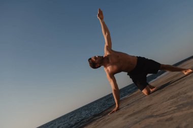 Yakışıklı, üstsüz adam arka planda deniz varken dışarıda yoga yapıyor.