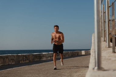 Rıhtımda koşan kendinden emin erkek sporcu. Arka planda deniz var.