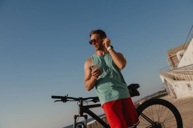 Bisiklete binen ve deniz kenarında telefon kullanırken kulaklık takan yakışıklı bir genç.