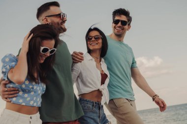 Kumsalda yürürken gülümseyen ve kaynaşan bir grup neşeli genç.