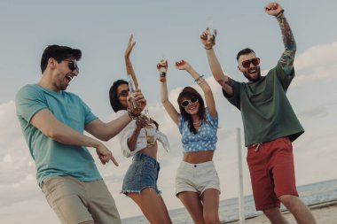 Güzel genç insanlar kumsalda dans ederken soğuk bira içip gülümsüyorlar.
