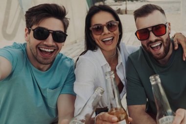 Kumsalda bira içerken selfie çeken dört mutlu genç insan.