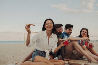 Mutlu genç arkadaşlar birlikte sahilde eğlenirken pizza ve içkinin tadını çıkarıyorlar.