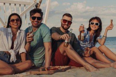 Kumsalda pizza ve biranın tadını çıkarırken kameraya bakan dört mutlu genç arkadaş.
