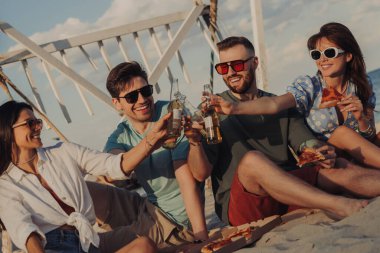 Bir grup neşeli genç arkadaş kumsalda pizzanın tadını çıkarırken birayla kadeh kaldırıyor.