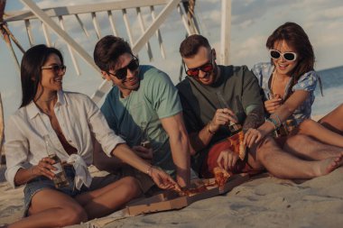 Bir grup mutlu genç arkadaş deniz kenarında vakit geçirirken pizza ve biranın tadını çıkarıyorlar.
