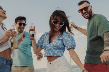 Bir grup güzel genç insan sahilde dans ederken bira içip gülümsüyor.