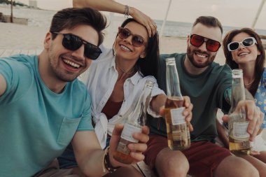 Birlikte sahilde bira içerken selfie çeken bir grup mutlu genç.