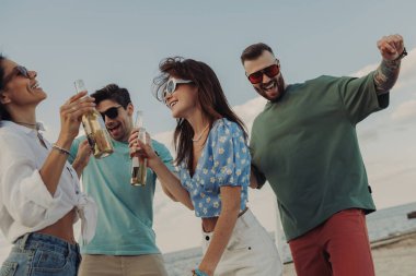 Bir grup güzel genç insan sahilde dans ederken bira içip gülümsüyor.