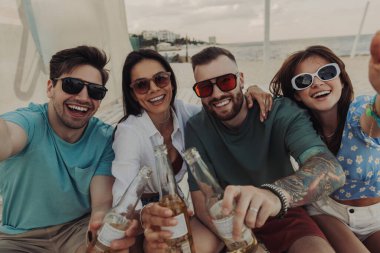 Sahilde bira içerken selfie çeken dört neşeli genç insan.