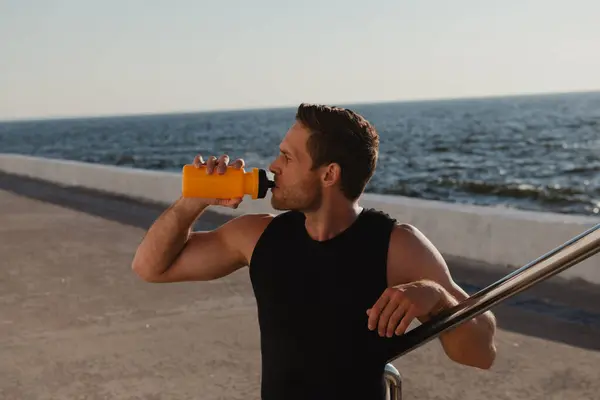 Handsome male athlete drinking water while resting after morning sport training seaside
