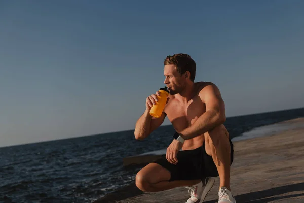 Handsome male athlete drinking water and enjoying sea view after morning training outdoors