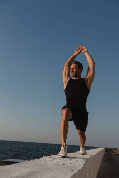 Уверенный мускулистый человек делает утренние упражнения на открытом воздухе с видом на море на заднем плане