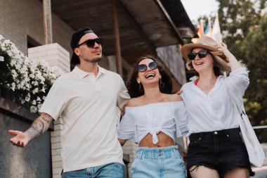 Üç neşeli genç arkadaş sokakta yürürken kucaklaşıp gülümsüyor.