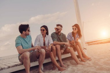 Kumsalda kaygısızca vakit geçirirken soğuk biranın tadını çıkaran dört mutlu genç insan.