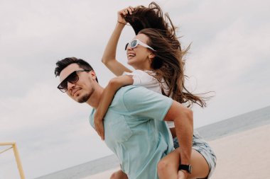 Yakışıklı genç adam kumsalda eğlenirken mutlu kız arkadaşını sırtında taşıyor.