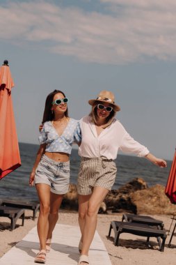 Kumsalda yürürken kucaklaşan ve gülümseyen iki çekici genç kadın.