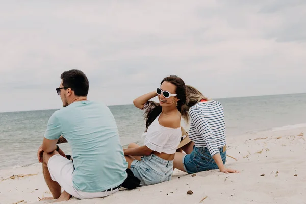 3人の美しい若者が砂の上に座り 一緒にビーチで無駄な時間を過ごしています — ストック写真